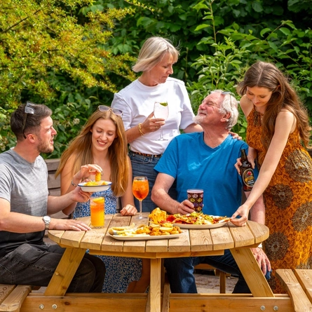 A mixed group of people seated around a wooden table, in a beer garden, enjoying a variety of sharers and drinks.