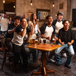 A group of people sitting or standing around a table in a pub, cheering while watching sports.