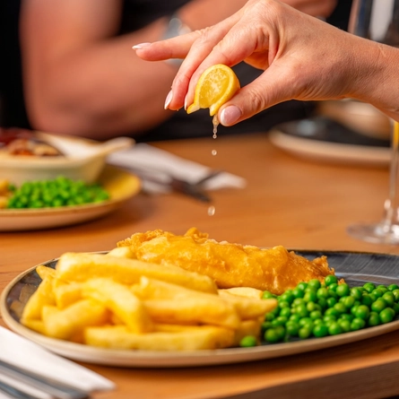 A hand squeezing a lemon wedge over a plated portion of fish and chips, at an indoor table.