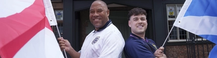John Barnes and Mark Thorburn standing back to back, outside in front of The Endbutt in Liverpool. John is wearing an England football shirt and holding the English flag, Mark is wearing a Scotland shirt and holding the Scottish flag.