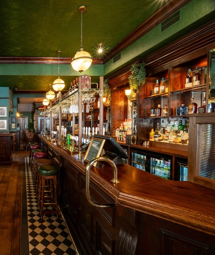 The bar and interior restaurant seating area at the King's Head, with bar stool seating; green, textured wallpaper on the ceiling; and red velvet curtains.