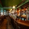 The bar and interior restaurant seating area at the King's Head, with bar stool seating; green, textured wallpaper on the ceiling; and red velvet curtains.