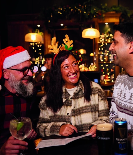 Three people are standing at a tall table with drinks,  in a festive, warmly lit bar. Two of them are wearing Christmas themed headwear.  The background features Christmas decorations, including garlands and lights, as well as hanging glasses above the bar area.