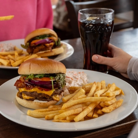 A close up view of a burger with fries and coleslaw, served on a plate which sits on a wooden restaurant table along with a glass of fizzy drink, and another plate of burger, fries, and coleslaw. A person sitting at the table is holding one of the fries, and a hand is holding the glass of fizzy drink.