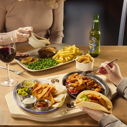 Two people sitting at a table, one of them is pouring sauce over a plate of steak, fries, peas, and onion rings, and the other person is using a fork to fill a fajita with food from the plates of meat, vegetables, nachos and sauce on the wooden board in front of them. A ramekin of onion rings, a glass of red wine and a bottle of Heineken 0.0% alcohol beer also sit on the table.