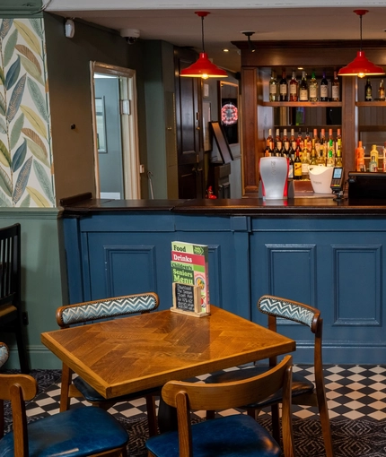A close up view of the blue, wood panelled bar at the Devonshire Arms in Sheffield, with restaurant seating, checkerboard patterned floor, and bottles of wine and alcoholic spirits on wooden shelves behind the counter.