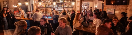 A crowded pub with wooden tables, warm lighting, and a busy bar lined with bottles and chalkboard menus at The Wilmington.