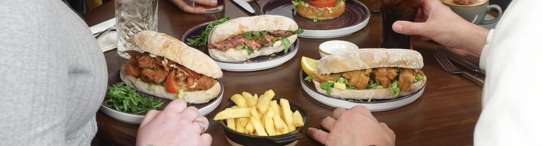 A group of four people seated around a small round wooden table, sharing a meal. The table is filled with plates of food, including baguette-style sandwiches, a burger, and several small bowls of chips. There are also drinks on the table, including glasses of soda with straws and a cup of coffee. Only the individuals’ torsos and arms are visible as they sit closely together, reaching toward the food.