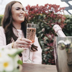 An image focusing on a glass of Rose Prosecco served in a champagne flute being enjoyed by a lady stood within the exterior beer garden seating area at a Crafted venue.