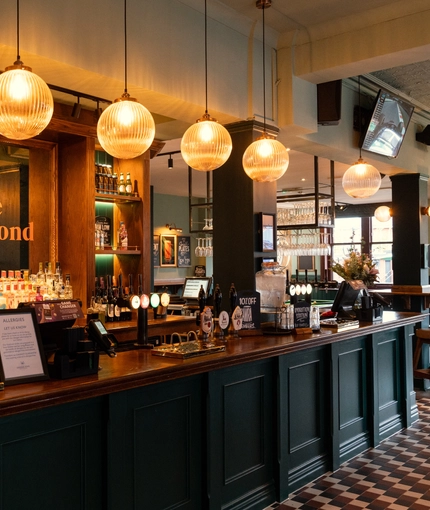 A close up view of the bar inside The Richmond Arms in Bournemouth, with spherical lights hanging above the counter; bottles of wine and alcoholic spirits on wooden shelves behind the bar; and a TV on the wall above.
