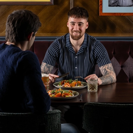 Two people sit across from each other at a wooden restaurant table. Each person has a plate of food in front of them featuring a flatbread topped with vegetables and garnishes. Drinks on the table include a pint of Level Head and a glass with ice.