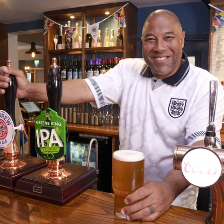 John Barnes behind the bar at The Endbutt in Liverpool, wearing an England football shirt and placing a pint of beer onto the counter.