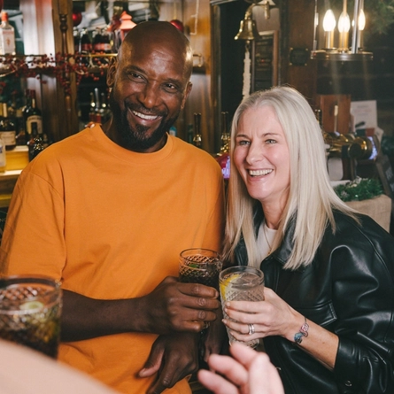 Three people next to the bar inside a pub, each of them holding a glass of drink.
