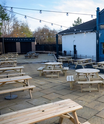 The exterior facade, signage, and paved beer garden seating area of The Kittoch in East Kilbride, with wooden picnic tables, and string lights overhead.