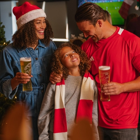 A festive pub setting decorated for Christmas, featuring a lit Christmas tree with red baubles and ribbon. Three people stand together beside the tree, including one wearing a red-and-white Santa hat and another wearing a red sports shirt. Two of the people are holding pints of beer, while a child wearing a red-and-white scarf stands between them. In the background, a football match is shown on a television screen and additional people wearing festive hats are gathered nearby.