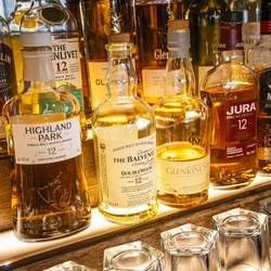 A close up view of bottles of different brands of whisky including Tamnavulin, Glenlivet, Glenkinchie, and Jura, standing on illuminated shelves inside a pub.