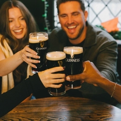 A group of friends sat round a table doing a cheers with pints of Guinness at a Greene King venue.