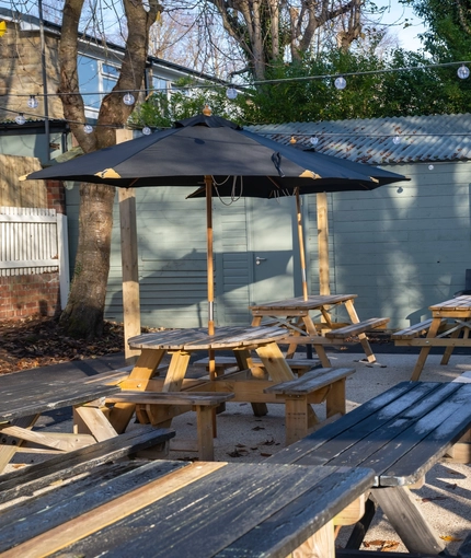 The exterior beer garden seating area at The Horsforth, with wooden picnic tables, a TV on the wall, shade umbrellas on some of the tables, and string lights overhead.
