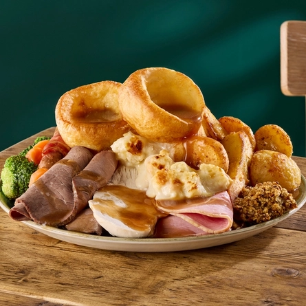 A close up view of a Standard Carvery featuring Yorkshire puddings, roast potatoes, vegetables, stuffing, roast meats, pigs in blankets, and gravy; all served on a plate which sits on a wooden table.