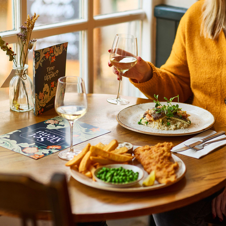 An image of a customer sat within the interior restaurant seating area enjoying a glass of white wine with 2 Main dishes and a glass of white wine sat on the table from the Set menu at a Chef & Brewer venue.