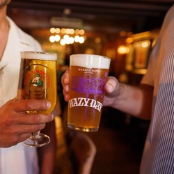 Two hands holding a pint of Hazy Day and Birra Moretti in an indoor bar setting.