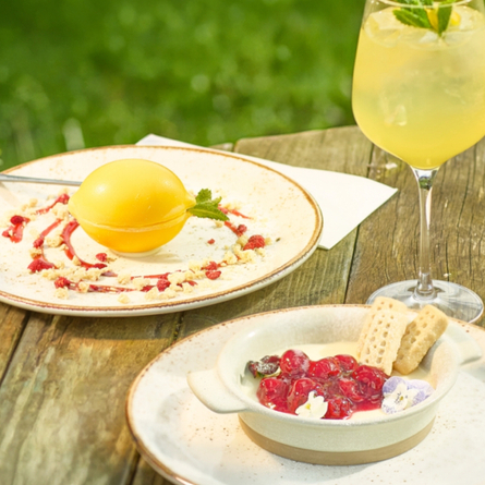 A banner showing a Lemon Cheesecake Bomb and a Lemon Posset dessert sat on a table with a cocktail style drink within an exterior beer garden seating area to advertise the new menu at Chef & Brewer venues.