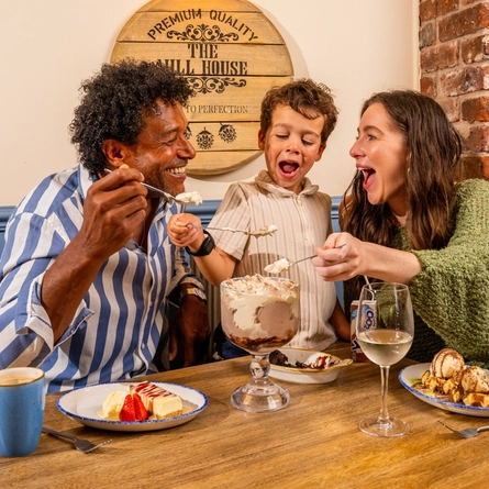 Two adults and a child are seated around a table in a pub, sharing a selection of desserts. The table features a large ice cream sundae, waffles topped with ice cream and sauce, cheesecake with strawberries, a chocolate dessert, a glass of white wine, a blue mug of coffee and a children's juice carton. Exposed brickwork, blue panelled walls and a wooden barrel-style wall sign create a warm and inviting pub setting.