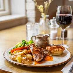 An advertising image of a plated Lamb Duo dish and glass of red wine sat on a table within the interior restaurant seating area at a Chef & Brewer venue.
