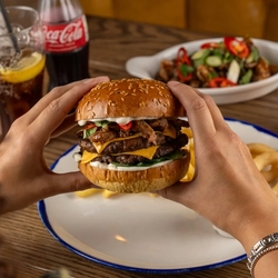 A pair of hands holding a Crispy Duck Burger over a plate on a wooden table.