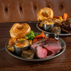 Two plates of roast dinner, including meat, Yorkshire pudding, vegetables and gravy, sitting on a wooden table.