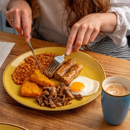 A close up view of an English Breakfast featuring sausages, fried egg, baked beans, hash browns, and mushrooms; served on a plate which sits on a wooden restaurant table. A serviette and a mug of coffee sit on either side of the plate, and a person at the table is about to pick up a hash brown with their knife and fork.