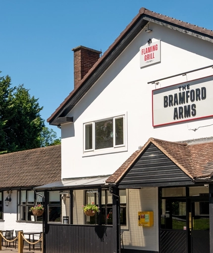 The exterior facade, signage, seating area, and car park of The Bramford Arms in Woodsetton, with accessible parking spaces near the entrance, and a ramp leading up to the double doors.