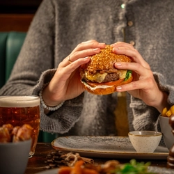 A person wearing a gray sweater is holding a sesame seed bun burger with both hands. The table in front of the person has a glass of beer, a small bowl of fries, two pepper grinders, and some other food items partially visible.