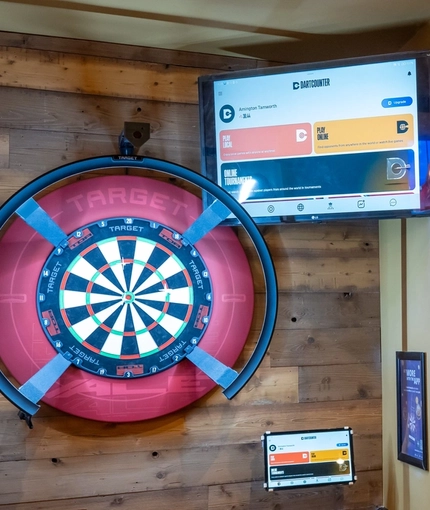 A close up view of the illuminated dartboard and TV on a wall inside The Amington. Black lettering on the yellow wall next to it reads "Let them fly!" in capital letters.