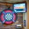 A close up view of the illuminated dartboard and TV on a wall inside The Amington. Black lettering on the yellow wall next to it reads "Let them fly!" in capital letters.