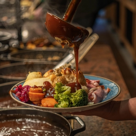 A close up view of a person at a carvery counter, holding a plate of roast dinner and using a ladle to pour gravy onto it.