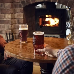 Two pints of Greene King IPA sitting on a table in front of a fireplace, with a man's hand around one pint.