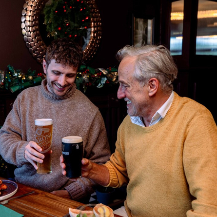 An image focusing on 2 people sat with others at a table within the interior restaurant seating area celebrating with a cheers while food dishes from the Festive Fayre menu at a Community Social venue.