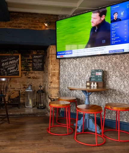 An interior restaurant seating area at The Royal Oak; with wooden flooring, a log burner in a large stone fireplace, and a TV on the wall.