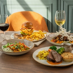 A selection of main meals from the Spring Menu at Chef & Brewer venues, served in bowls or on plates and sitting on a wooden table along with two glasses of wine.