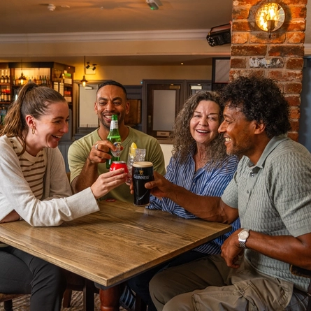 Four people gathered around a wooden table in a pub, raising assorted drinks in a toast. The drinks include a pint of Guinness, a bottled alcohol-free beer, a can of soft drink and a mixed drink with ice and lemon. The pub interior features exposed brickwork, a stocked bar, warm lighting and wooden furnishings.