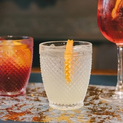 Three cocktail type drinks lined up on a bar, one with red liquid in a stemmed glass, one is a pale liquid in a lowball glass and the third is a red liquid in a lowball glass.  All have ice and a slice of fruit garnish.
