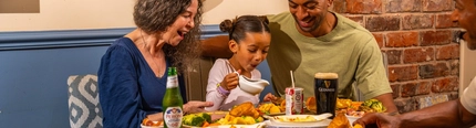 Three people seated around a wooden table in a pub, sharing Sunday roast meals. One person pours gravy from a small jug onto a plate of roast dinner with Yorkshire puddings, vegetables and potatoes. A bottle of alcohol-free beer, a pint of Guinness and a children's juice carton sit among the dishes. The background features blue panelled walls, exposed brickwork and a wooden barrel-style sign reading “The Mill House”.