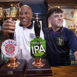 John Barnes and Mark Thorburn standing behind the bar at The Endbutt in Liverpool. John is holding one of the beer taps, and Mark is holding out a pint of beer.