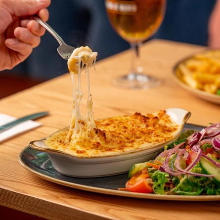 A hand holding a forkful of Mac & Cheese, over a roasting dish of Macaroni Cheese at an indoor table..