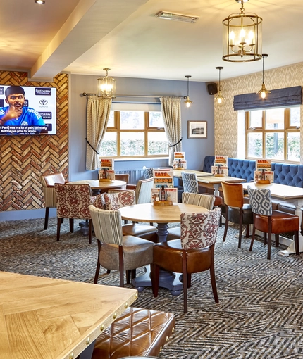The interior restaurant seating area of The Flying Horse in Heald Green, with wooden tables, upholstered chairs, and a TV on the wall.