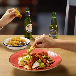 A bowl of nachos topped with sauce, salsa, guacamole, and sour cream. The bowl sits on a wooden table next to a small bowl of chicken strips, a bottle of Peroni lager, and a bottle of Heineken 0.0% alcohol beer.