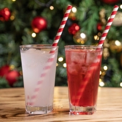 Two festive cocktails served in highball glasses with ice and candy cane pattern straws. The glasses sit on a wooden table and a Christmas tree is visible in the background. 
