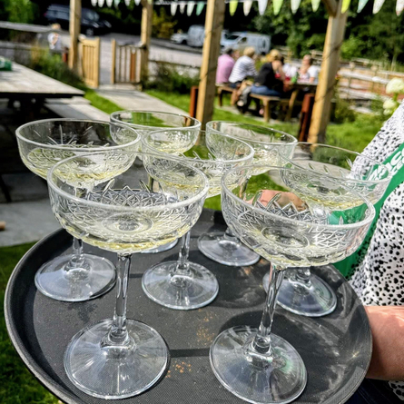 An image of a silver tray with glasses of sparkling white wine being carried through a beer garden to advertise a Bank Holiday Garden Party in aid of Macmillan at The Cart & Horses.