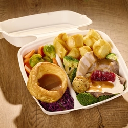 A close up view of a takeaway box filled with Yorkshire pudding, roast potatoes, broccoli, carrots, Brussels sprouts, stuffing, slices of roast meat, cranberry sauce, and gravy; sitting on a wooden table.
